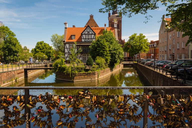 Scenic view on half-timbered building on the river island in european city, church tower on background and padlocks on bridge of love on foreground. Miller house, Grand Mill, Old Town, Gdansk, Poland